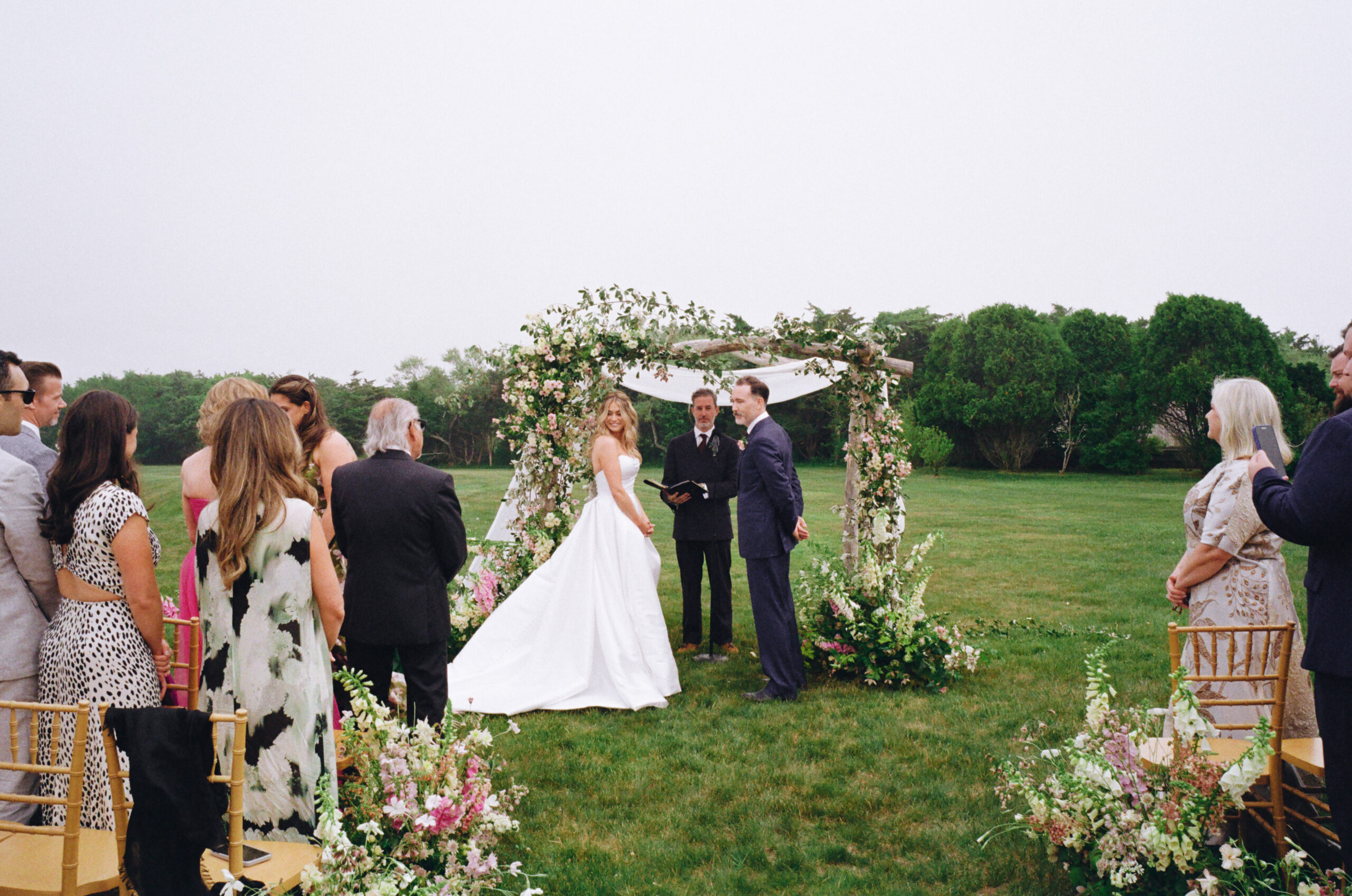 Couple standing under flower arch with officiant during outdoor wedding ceremony in Massachusetts.