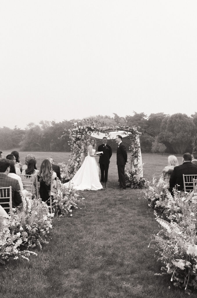 Black and white film wedding photo of couple exchanging vows beneath floral arch.