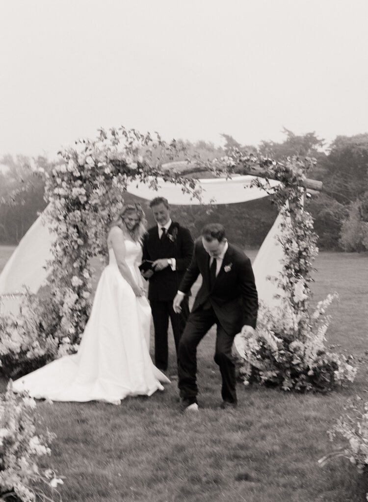 Couple breaking glass during outdoor wedding ceremony – candid black and white film wedding moment.