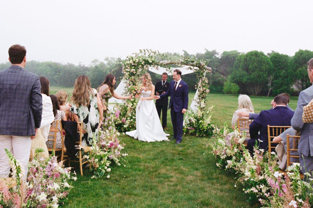 Bride and groom smile as they walk hand in hand down the aisle lined with floral arrangements, pausing to greet guests.