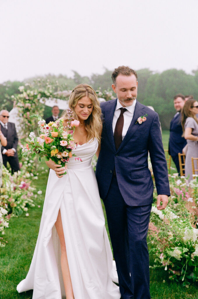 Bride and groom walk arm in arm, looking down as they smile, with lush floral decor behind them.