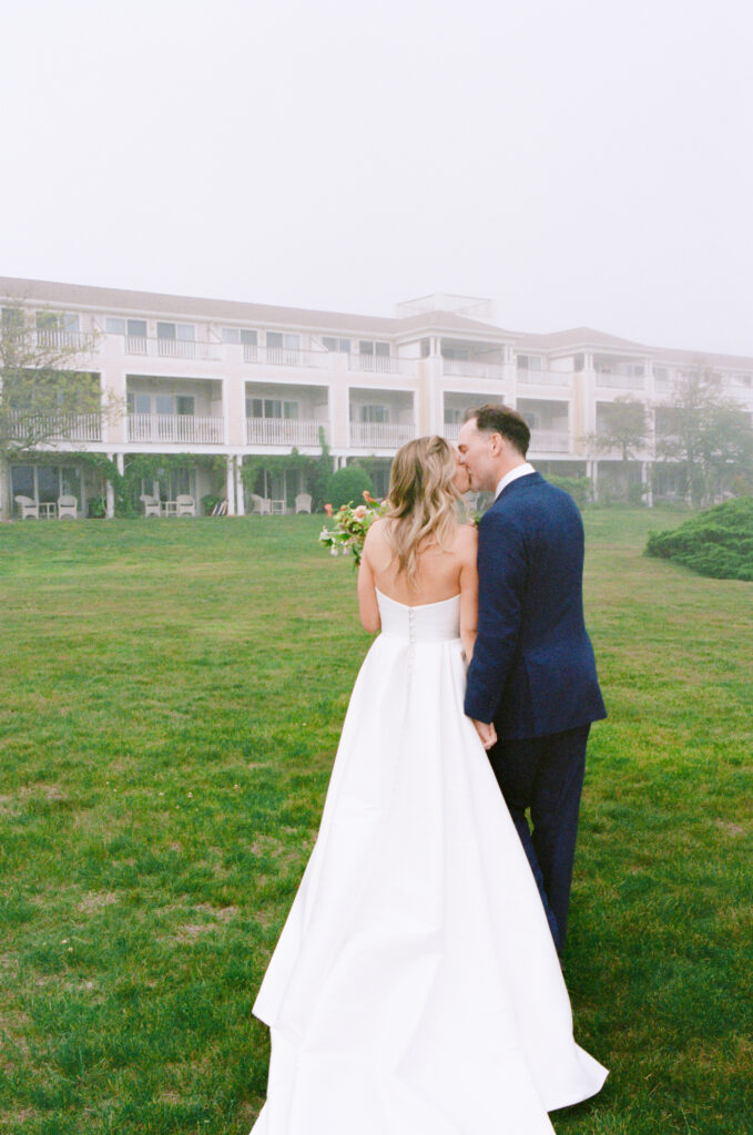 Couple walks away from the ceremony, kissing in front of a misty white building in the background.