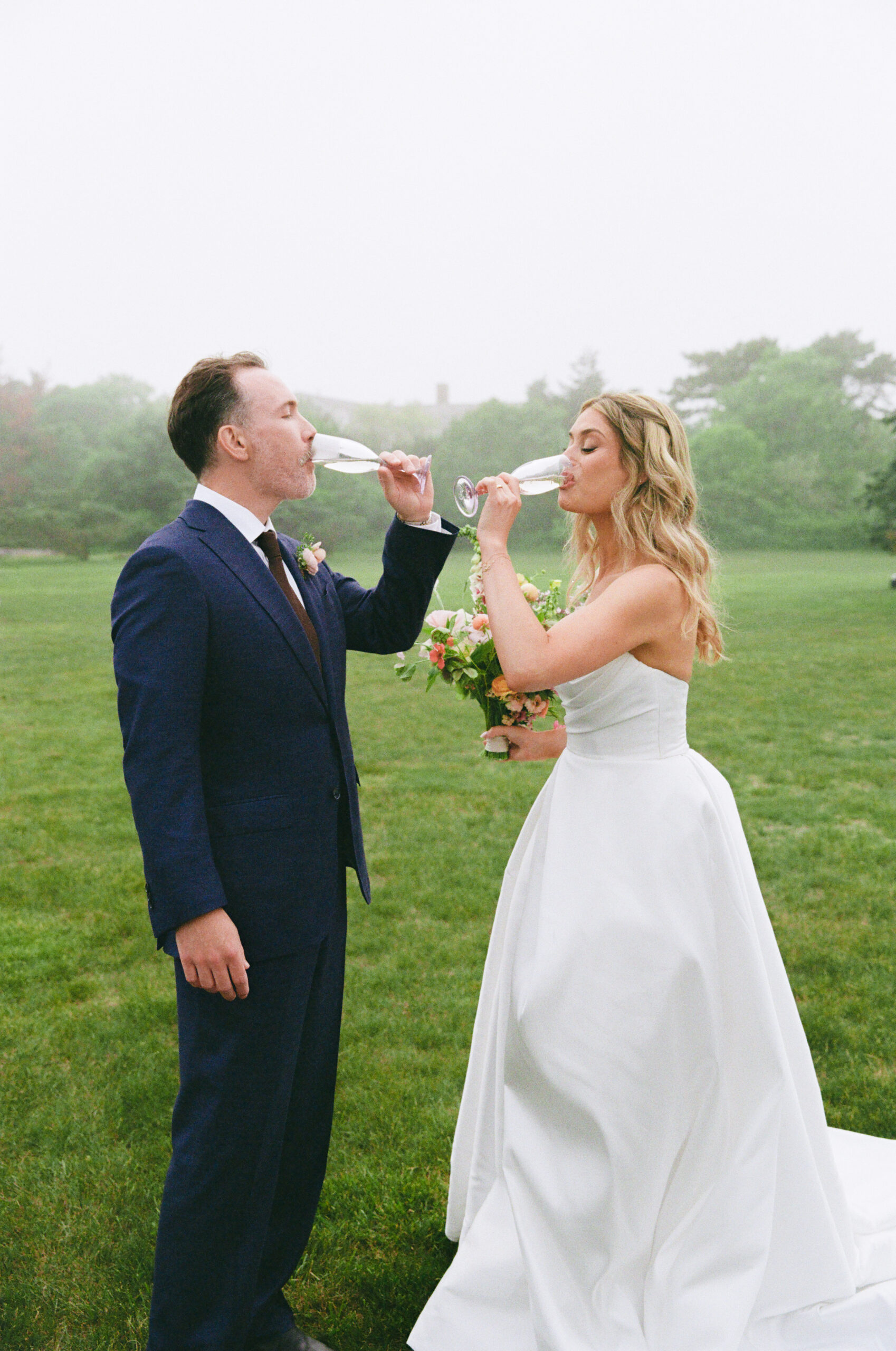 Bride and groom toast with champagne flutes on the lawn, the bride holding her bouquet in her other hand.