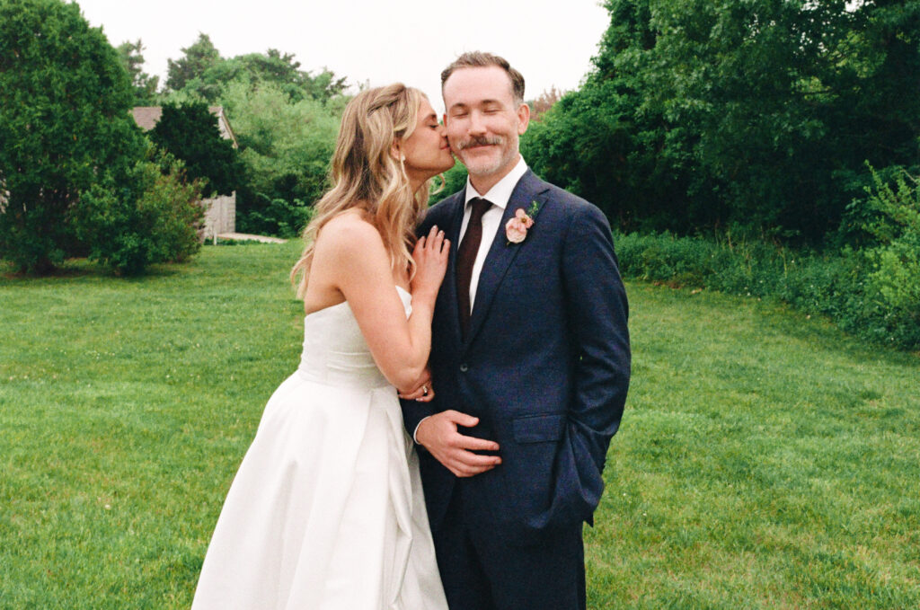 Romantic color film portrait of the bride and groom at their Martha’s Vineyard wedding, standing in a foggy field with trees in the background. The bride wraps her arms around the groom, nestling her nose against his cheek as he smiles with one hand in his pocket, creating a timeless and scenic wedding photo.