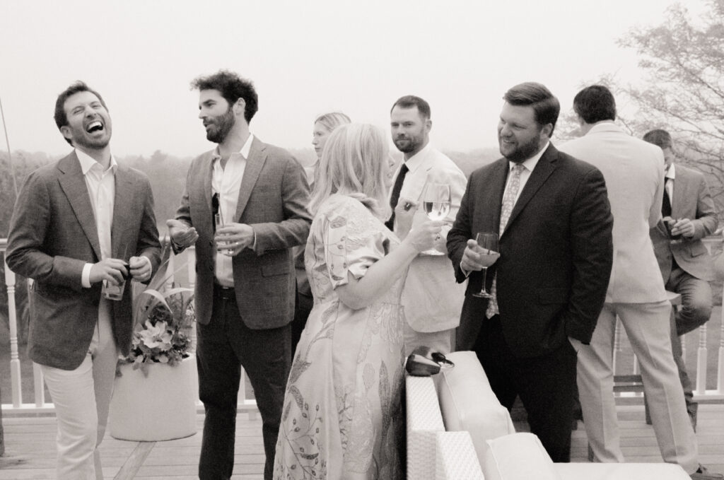 Wedding guests mingling and laughing with drinks on outdoor deck, candid film wedding photography in New England