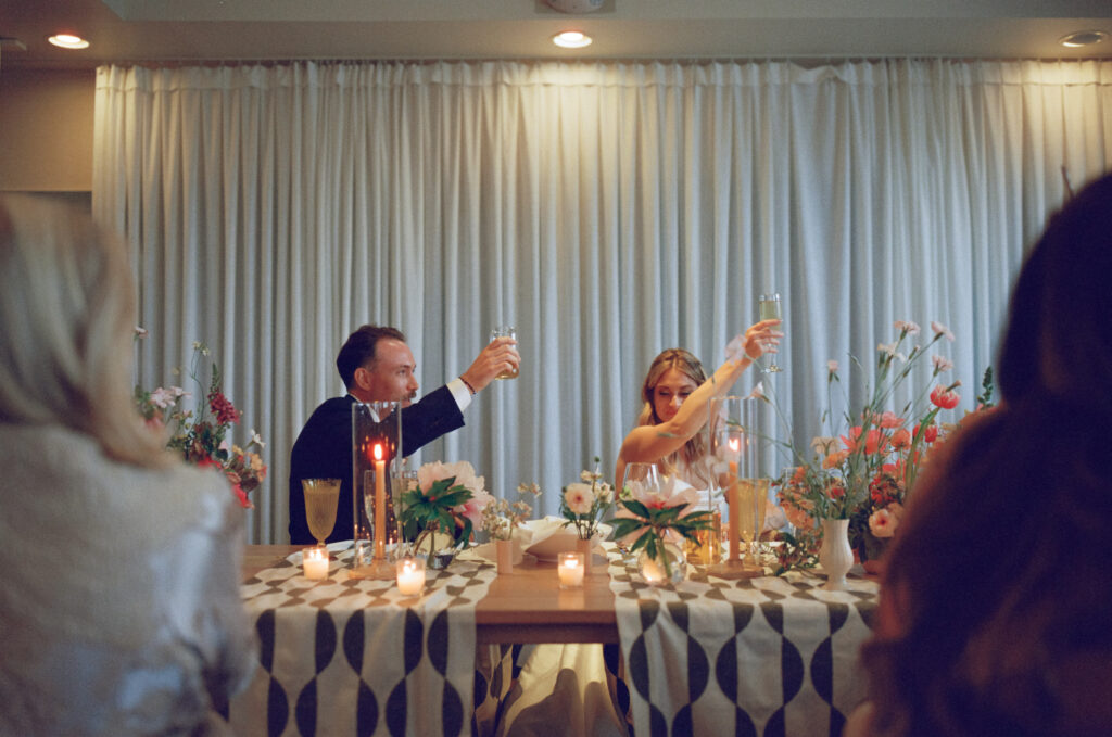 A man and woman seated at a decorated table raise glasses for a toast, surrounded by candles, flowers, and other guests.