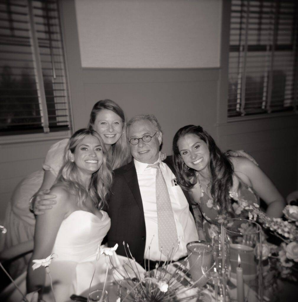 Candid black and white Holga 120 medium format film photo of bride Olivia sitting with her sister, uncle, and cousin during the Martha’s Vineyard wedding reception. Elegant, timeless group portrait at the dinner table.