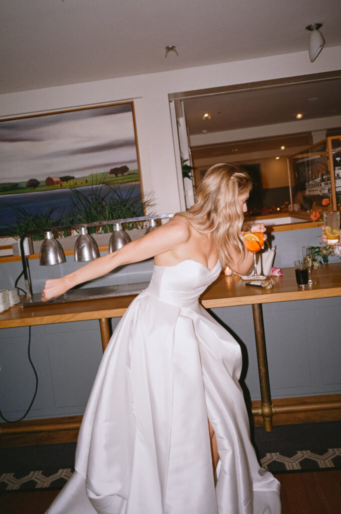 “Candid 35mm color film flash photo of the bride on the dance floor at her Martha’s Vineyard wedding, sipping an Aperol Spritz mid-shuffle, captured in a fun in-between moment.