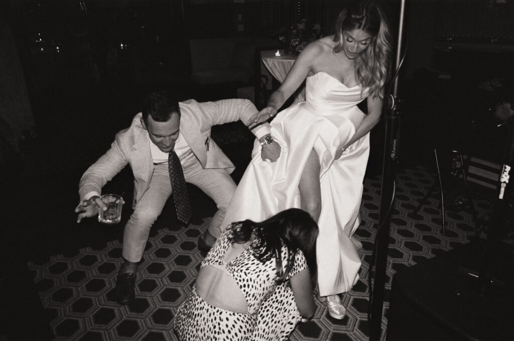 Black and white 35mm film flash photo of bride Olivia on Martha’s Vineyard lifting her wedding dress to remove her heels, with her best friend and husband helping her before the first dances.