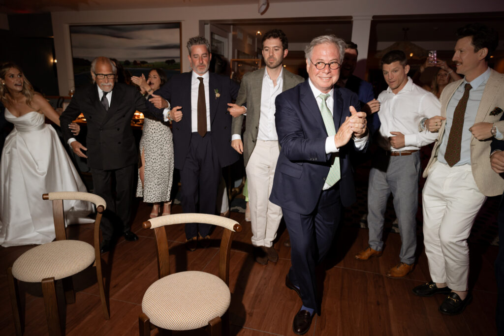 Color flash candid photo at a Martha’s Vineyard wedding showing the bride’s uncle setting down chairs before the Hora as guests circle around holding hands with big energy.