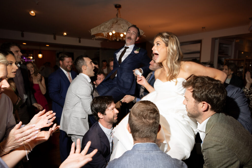Candid color photo of the bride and groom lifted on chairs during the hora at their Martha’s Vineyard wedding reception. Captured with flash, the image is full of movement, energy, and joy as guests cheer and celebrate.