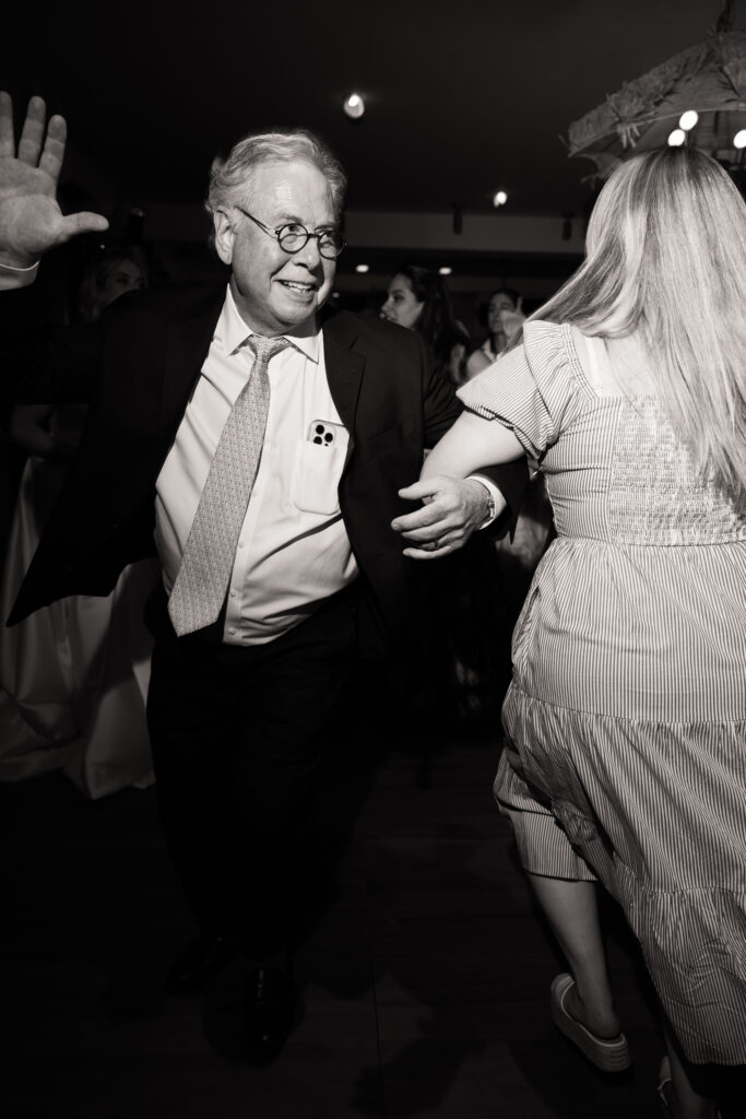 Black and white flash digital photo of guests dancing during the Hora at a Martha’s Vineyard wedding, with an uncle and his daughter locking arms and raising their hands in joyful celebration.