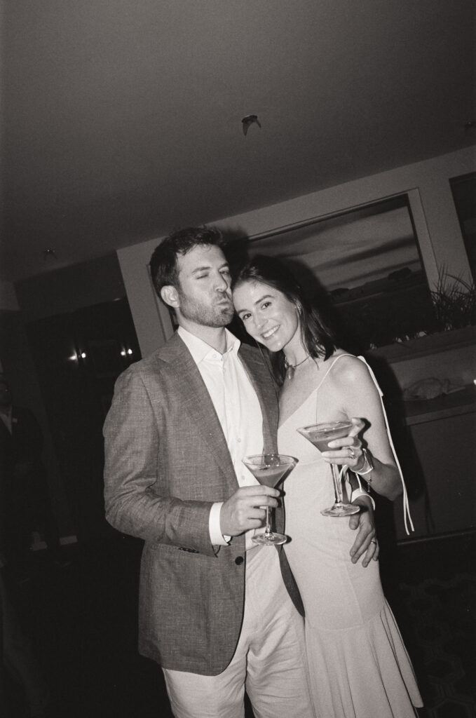 Black and white 35mm film flash photo of wedding guests at a Martha’s Vineyard reception, with one holding a martini glass and another playfully making a kissy face toward the camera, a candid yet editorial-style moment of fine art film wedding photography