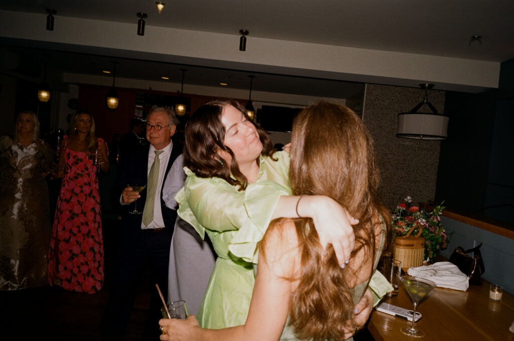 Candid 35mm color film flash photo of two wedding guests dancing together during a Martha’s Vineyard wedding reception, with one in a green dress playfully throwing her arms over her friend.