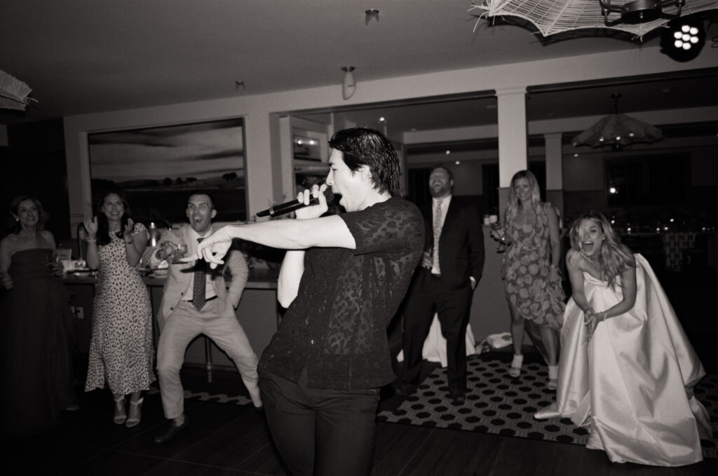 Black and white 35mm film flash photo of the lead singer performing on the dance floor during a Martha’s Vineyard wedding reception, energetically pointing toward the crowd with guests cheering in the background.