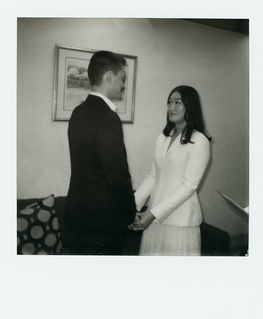 The bride and groom interlock hands at their ceremony, black and white Polaroid photograph.
