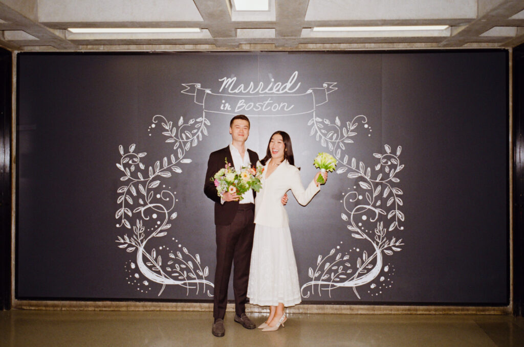 The bride and groom hold floral bouquets in front of "Married in Boston" sign at Boston City Hall. 