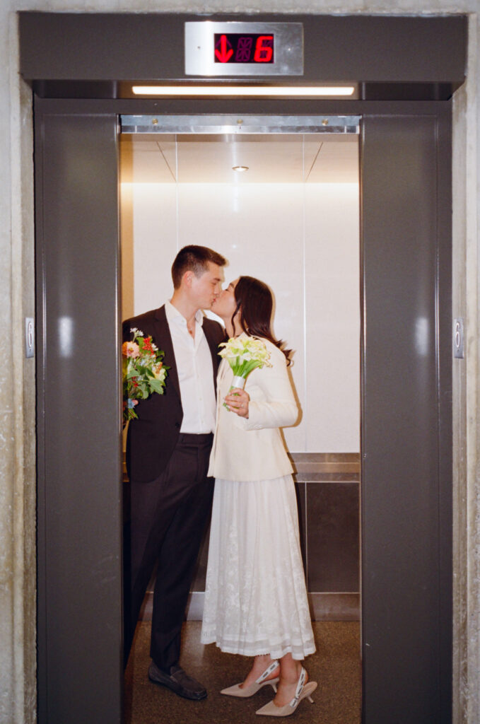 The bride and groom kiss, holding bouquets, as the elevator door shuts, documentary-style Boston City Hall film wedding photography.