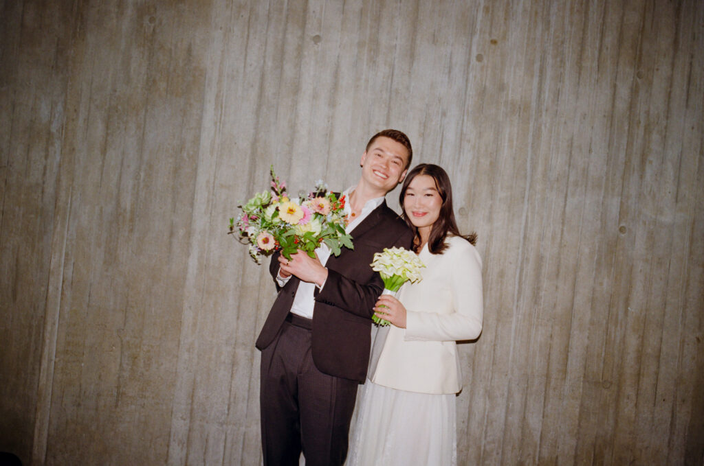 Bride and groom smile and pose at the camera against the backdrop of Boston City Hall. The groom holds a lush bouquet, film photography by Castillo Holliday Photo + Film