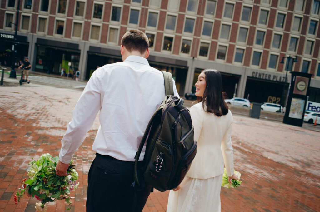 The groom and bride walk ahead towards Center Plaza with bouquets in hand, after it rains, 35mm photograph.