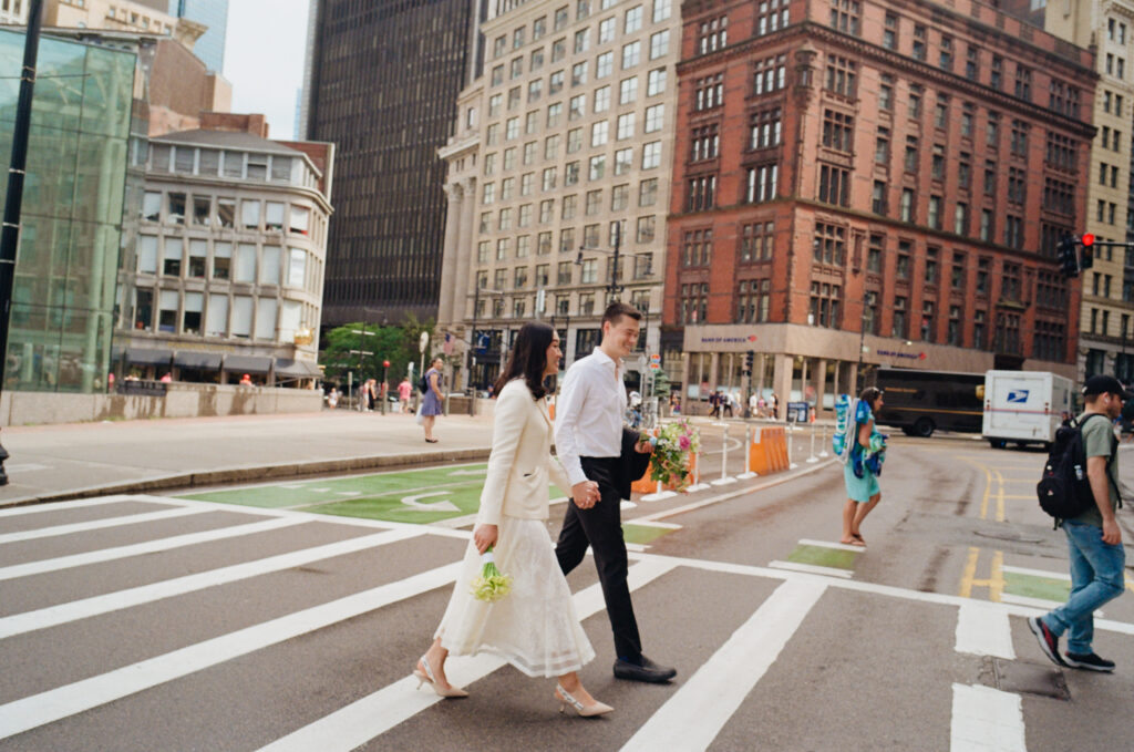 The bride and groom hold their bouquets as they walk across the street in bustling downtown Boston after departing from their Boston City Hall wedding.