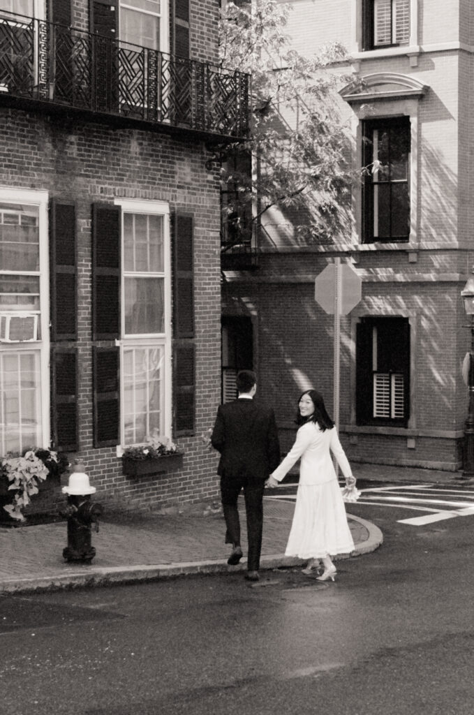 Bride and groom hold hands while walking around a corner in Boston's Beacon Hill neighborhood, smiling -- candid black and white film wedding photo