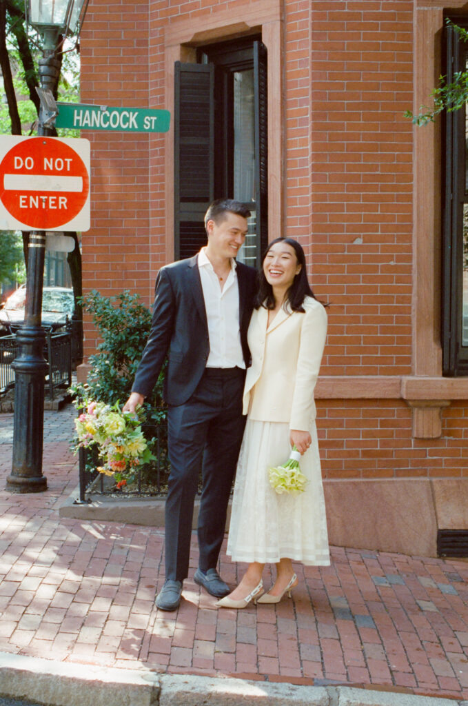Bride and groom laugh, facing the camera on corner in Beacon Hill in Boston. Bride and groom hold bouquets of lush florals -- documentary 35mm wedding photograph.