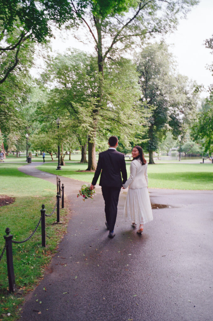 Bride and groom walk the Boston Public Garden after it rained, looking back at the camera, amongst the lush green trees.