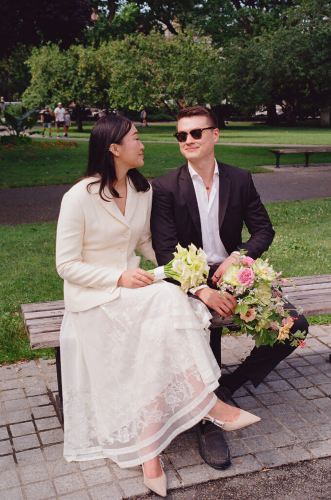 The bride and groom sit on a bench in the Boston Public Garden, looking at each other, with white and pastel blooms. 