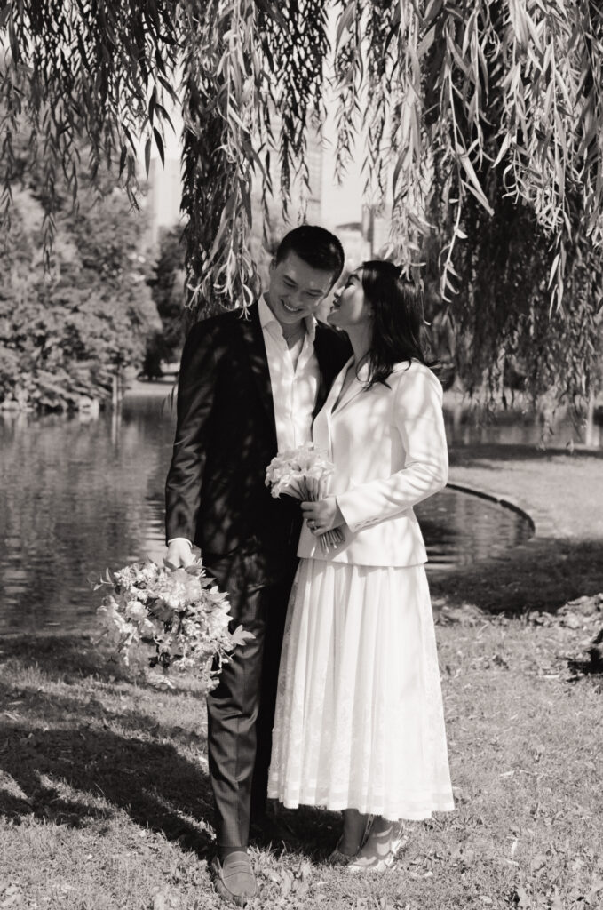 Black and white photograph of the bride whispering into groom's ear under a willow tree in the Boston Public Garden.