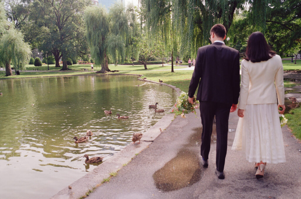 Bride and groom walk together along the Boston Public Garden pond, looking at the ducks, with bouquets in hand.