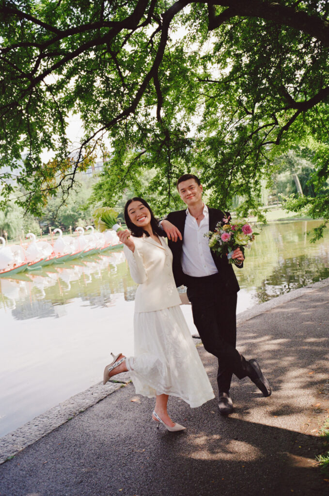 Bride and groom hold up their bouquets in front of the Boston Public Garden swan boats, the bride kicking her foot in the air after finishing their Boston City Hall elopement.
