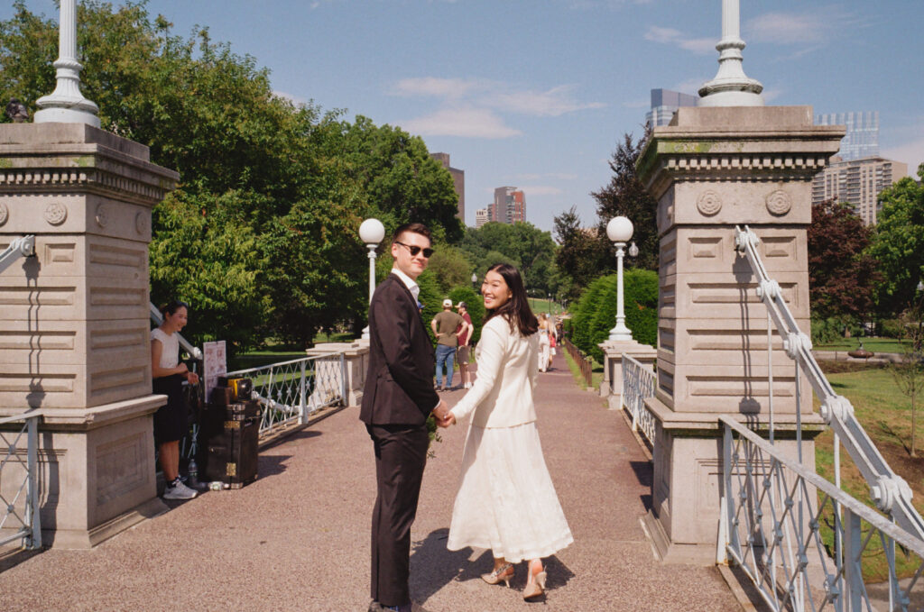 Bride and groom look back at the camera while crossing the bridge in the Boston Public Garden, laughing --- candid 35mm film wedding photograph.