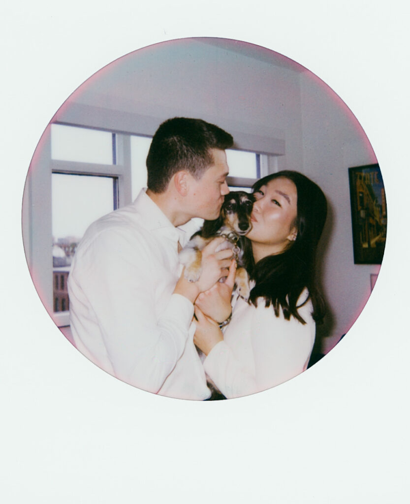 The bride and groom hold up their dachshund between themselves, giving it kisses-- shot on Polaroid in Boston