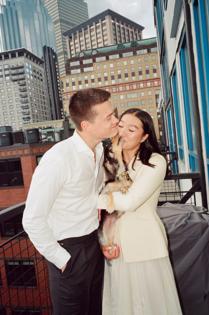 The dog, held in the bride and groom's arms, kisses them, backdropped by the Boston skyline.
