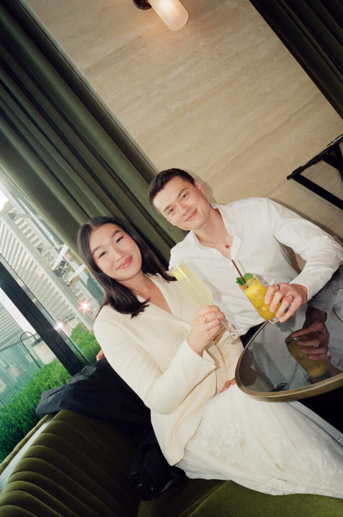 The bride and groom sit together facing the camera with tall, yellow drinks, after finishing their Boston City Hall elopement. 