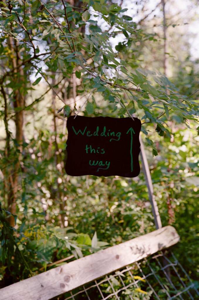 A black "Wedding this way" sign hung on a tree branch as a ceremony detail for an elopement day, shot on color film.