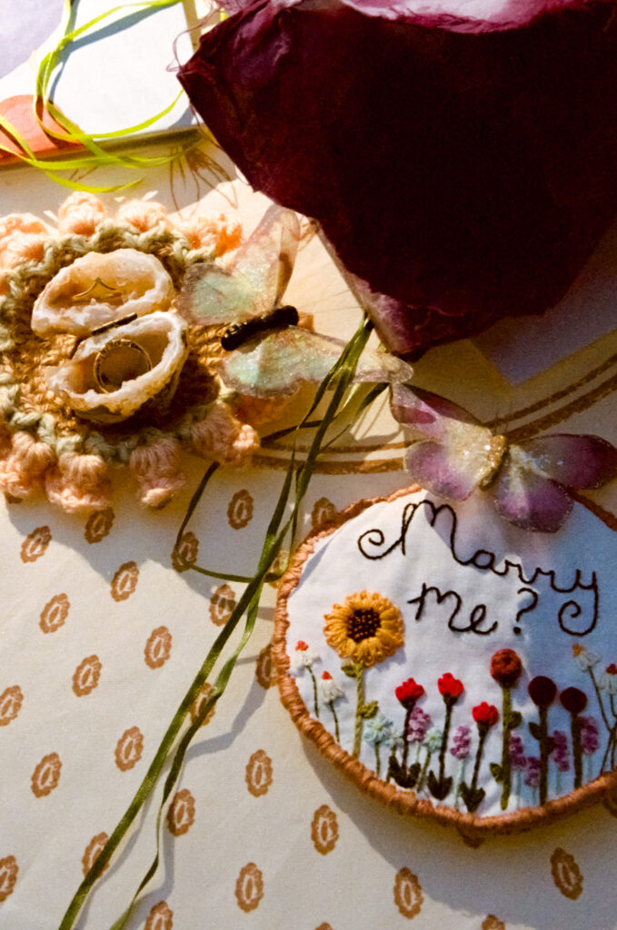 Detail close-up of various relationship-trinkets, including rings and ring boxes, a circular embroidered "marry me" sign, and scattered fake butterflies