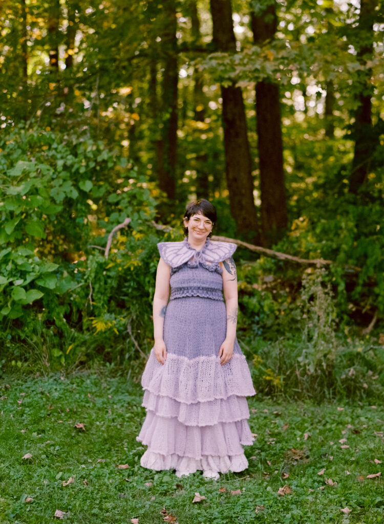 A color film photograph of a person in her elopement-day dress, hand-crocheted with purple yarn amongst a bright green forest backdrop. 
