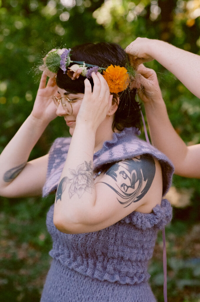 Bride holds her flower crown in place as hands behind her tie it.