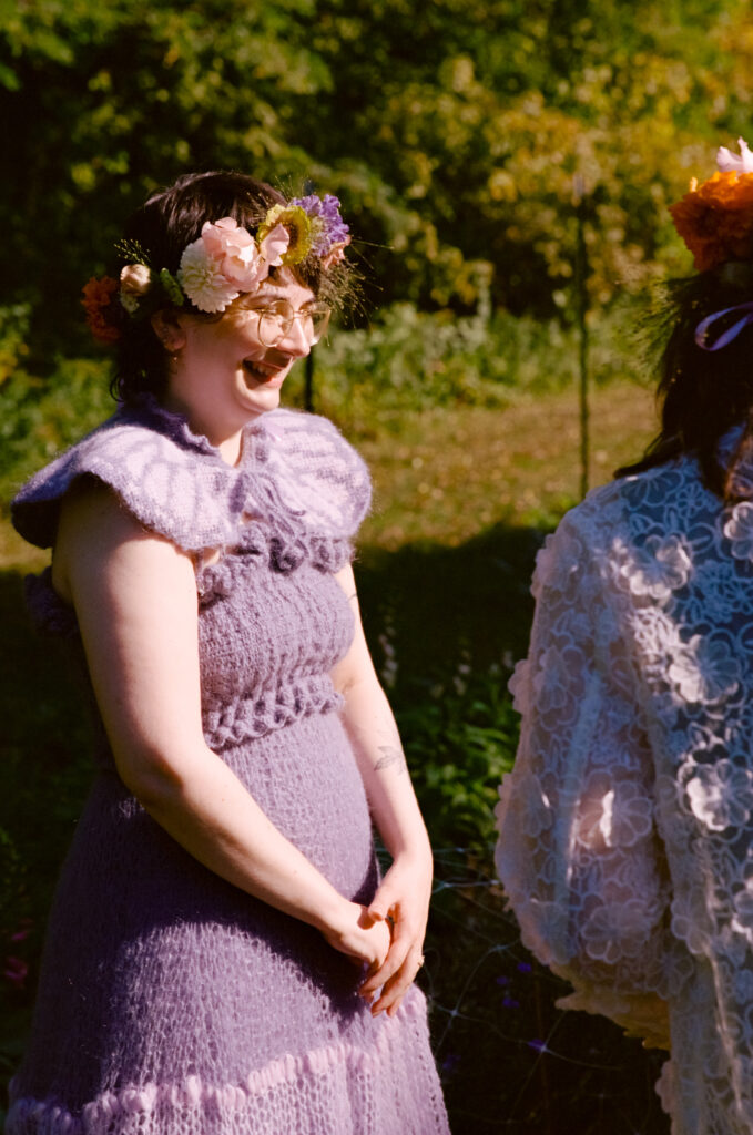 Bride gives a big smile in a close-up shot at her wedding alter, dressed in a purple crocheted dress and a flower crown.