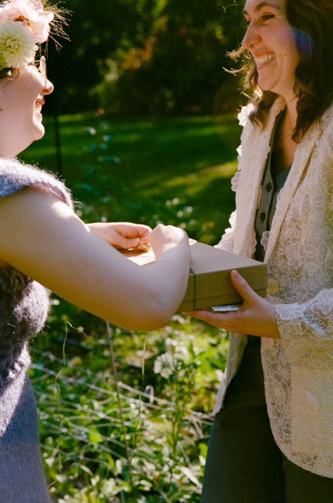 A bride holds a gift box out to her partner, who is unrapping the string as the smile bright at one another. 