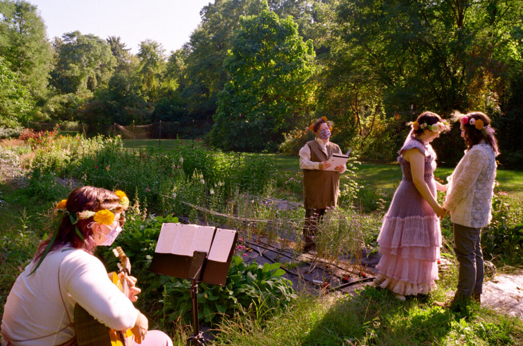 A wide angle photograph of an LGBT wedding ceremony, picturing the couple, the officiant, and a guitarist amongst a flower field.