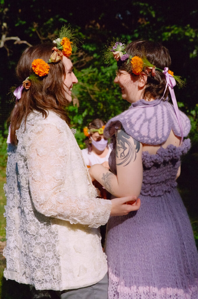 A couple gleams at each in other in their colorful wedding day attire, complete with orange flower crowns.