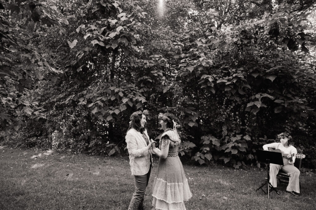 Black and white wide angle photograph of couple dancing in a field, accompanied by a musician. 