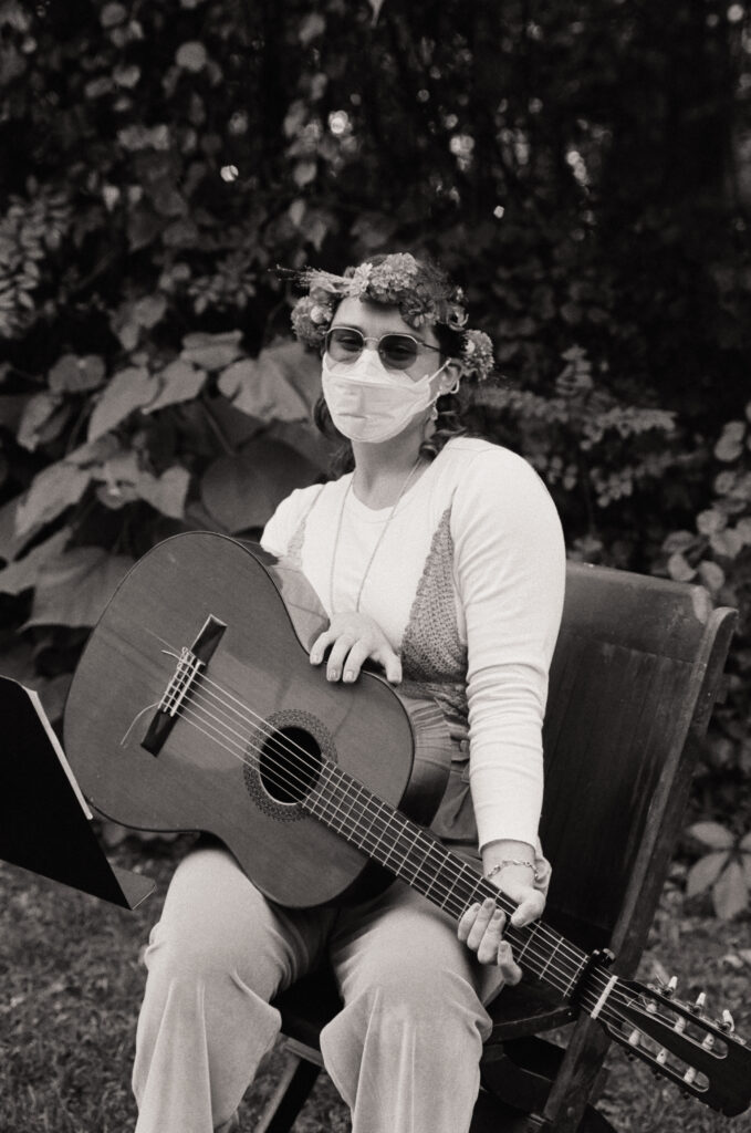 Portrait of a guitarist sitting. They hold their guitar on their lap and wear a flower crown in celebration of the wedding ceremony.