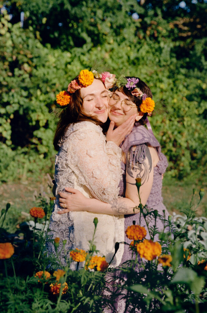 Intimate film portrait of newlywed couple grasping each other. Eyes closed with smiles, they bask in the sun amongst marigold flowers.