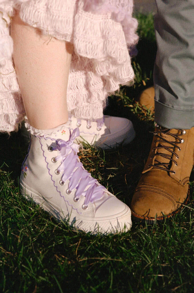 Detail photograph of both brides' shoes, on the left a pair of white and purple Converse and the right tan dress shoes. 