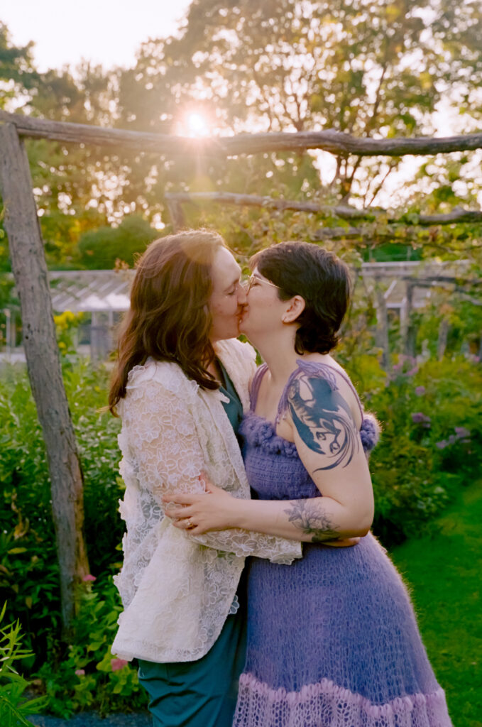 Couple shares kiss within a wooden archway, backdropped by a sun flare coming in overhead.