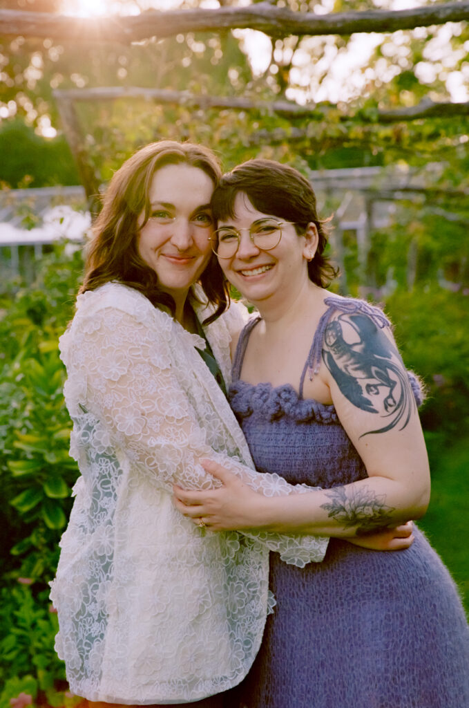 Couple looks forward with bright smiles, holding each other in a path of greenery.